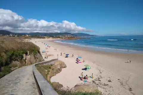 Un paseo de piedra bordea la **playa llas**, donde gente descansa bajo sombrillas y el mar azul invita a un baño.