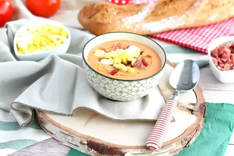 Schüssel mit Gazpacho, garniert mit Ei und Schinken. Daneben sind Tomaten, Brot und weitere Zutaten für kalte Tapas.
