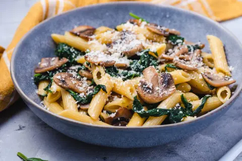 Penne z pieczarkami i szpinakiem, posypane parmezanem, w szarej misce.