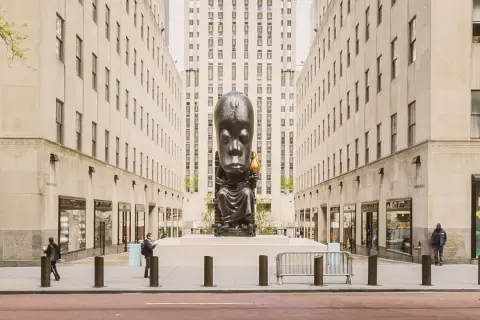 A monumental sculpture of a seated figure with a large head, holding a torch, stands as a striking example of public art between two imposing buildings.