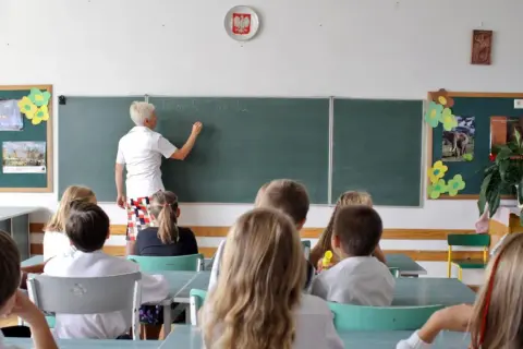 Nauczycielka pisze na tablicy, a uczniowie siedzą w ławkach. To scena z lekcji, która mogłaby się odbywać tuż przed tym, jak skończyło się gimnazjum w Polsce.