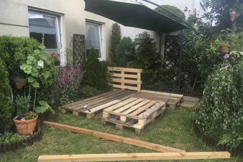 Taras z palet w trakcie budowy, otoczony zielenią i kwiatami. Pod dużym parasolem powstaje miejsce do relaksu.