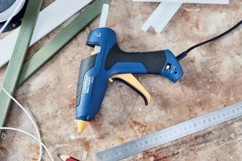 Pistolet à colle bleu et jaune, prêt à l'emploi pour apprendre comment coller du plastique. Des bâtons de colle et des règles sont à proximité.