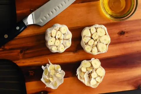 Na desce do krojenia leżą przekrojone główki czosnku, nóż i miseczka z oliwą. Czy czosnek obniża cholesterol?