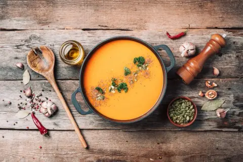 Pomarańczowa zupa dyniowa w garnku, z pestkami dyni, natką i przyprawami na drewnianym stole.