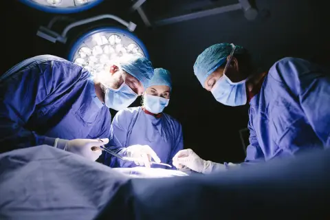 Trois médecins spécialistes en blouse bleue, masqués, concentrés sur une opération sous les lumières vives du bloc opératoire.