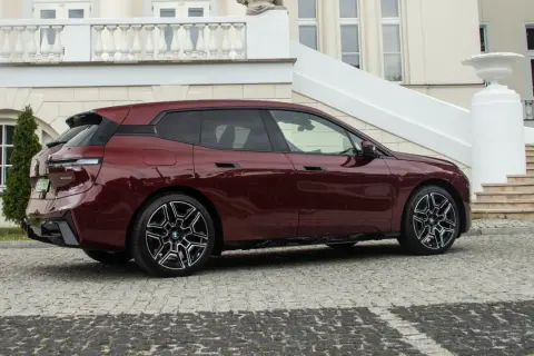Bordowy BMW iX z eleganckimi felgami stoi przed luksusową posiadłością z kamienną nawierzchnią.