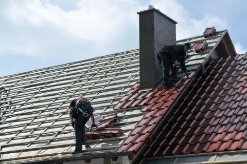Dwaj dekarze układają dachówki. Widać rusztowanie i komin. Trudno ocenić ile dachówek na m2.