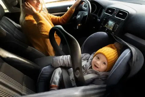 Uśmiechnięte dziecko w żółtej czapce w foteliku na przednim siedzeniu samochodu, obok kierowcy.