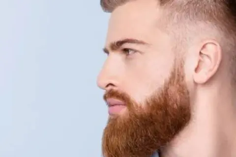 Un homme au profil droit, avec une barbe rousse soignée, tailler sa barbe en dégradé. Ses cheveux sont courts sur les côtés.