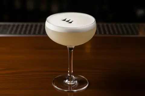 En elegant pisco sour med ett fluffigt skum och tre små mörka prickar på toppen, serverad i ett coupeglas på en träbar.