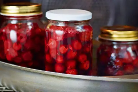 Drei Gläser mit eingelegten Kirschen stehen in einem Metallbehälter. Die roten Früchte schimmern im Saft.