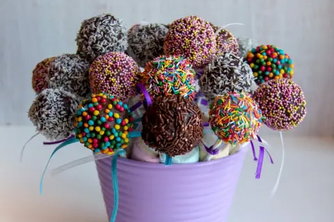 Kolorowe cake pops z jasnego biszkoptu, udekorowane posypkami i wiórkami kokosowymi, w fioletowym wazonie.