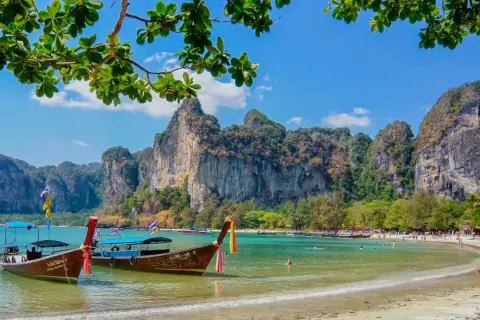 Två longtailbåtar guppar i det turkosa vattnet vid en vacker thailand strand. Imponerande klippor reser sig bakom den gröna djungeln.