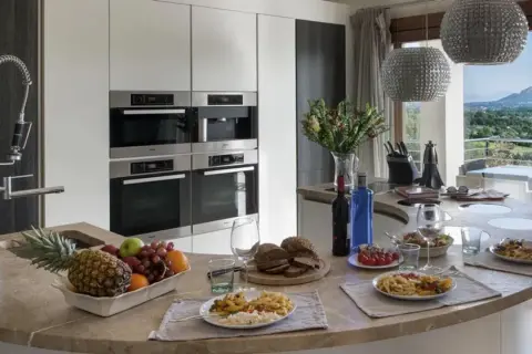 Cocina moderna con encimera curva, electrodomésticos integrados y vista al paisaje. Ideal para un contrato de alquiler temporal, con comida servida y frutas frescas.