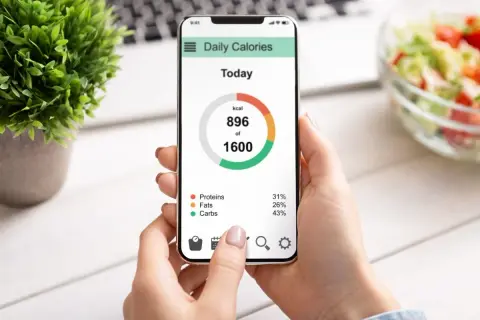 Dłoń trzyma smartfon z aplikacją "Daily Calories", pokazującą spożycie 896 z 1600 kcal. Widoczny jest wzór mifflina w rozkładzie białek, tłuszczów i węglowodanów.