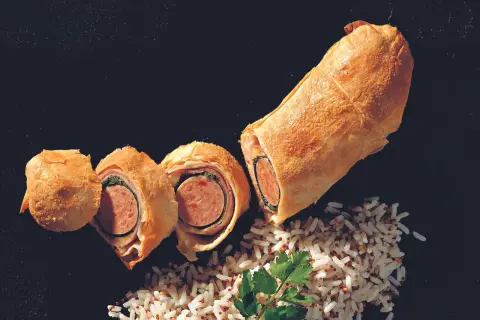 Herzhaft belegter Bratwurst im Teigmantel, aufgeschnitten und mit Reis garniert.