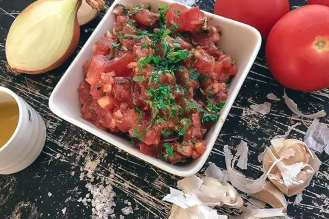 En skål med färsk tomatsalsa, redo för ett tomatsalsa recept. Ingredienser som lök, vitlök och tomater syns runt omkring.