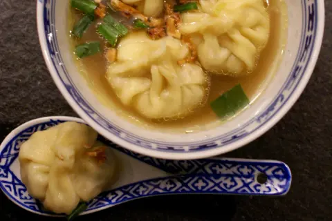 A miseczce z niebiesko-białym wzorem znajduje się aromatyczna zupa wonton z zieloną cebulką i chrupiącymi dodatkami. Na łyżce leży jeden pierożek wonton.