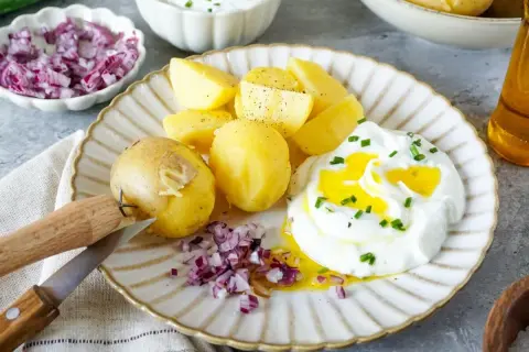 Kartoffeln mit Quark, garniert mit Schnittlauch und verfeinert mit Öl. Dazu gibt es gehackte rote Zwiebeln und ein Messer zum Schälen.