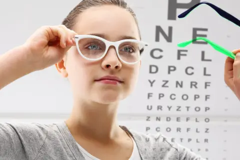 Dziewczynka z wadą wzroku minus 1 przymierza okulary na tle tablicy Snellena.