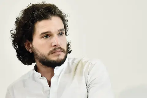 Hombre con pelo rizado oscuro y barba, viste camisa blanca.