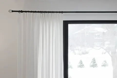Czarny karnisz z białymi zasłonami na tle okna ze śnieżnym krajobrazem.