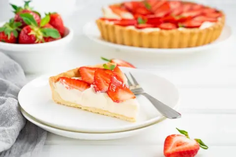 Pyszna tarta truskawkowa z kremowym nadzieniem i świeżymi owocami. Kawałek ciasta na talerzyku, obok cała tarta i miseczka truskawek.