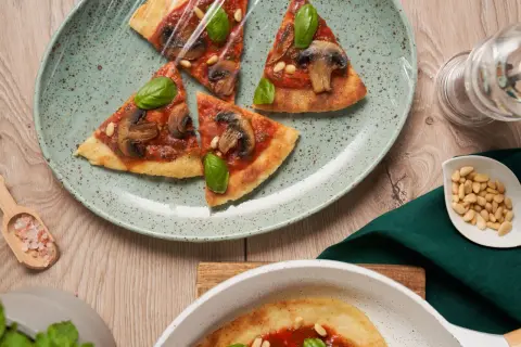 En tunn, krispig flatbread pizza med svamp och basilika, täckt med plastfolie. Pinjenötter och rosa salt ligger bredvid.
