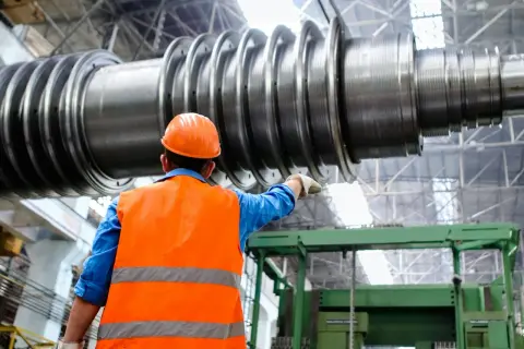 Pracownik w pomarańczowej kamizelce i kasku wskazuje na ogromny wał turbiny. To przykład precyzyjnej produkcji jednostkowej.