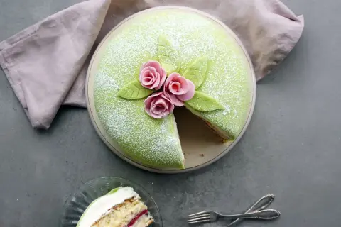 En vacker prinsesstårta med grönt marsipanlock, rosa rosor och blad. Ett stycke är uppskuret, perfekt för att inspireras av ett prinsesstårta recept.