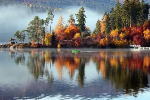 Ein idyllischer Urlaub im Oktober in Deutschland: Nebel steigt vom See auf, bunte Herbstbäume spiegeln sich im Wasser, eine grüne Ruderboot gleitet vorbei.