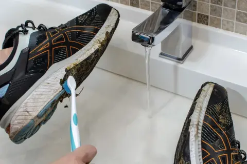Come lavare le scarpe da running: una mano con uno spazzolino pulisce il fango dalla suola di una scarpa sotto un rubinetto aperto.