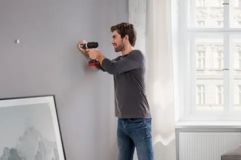 Uomo usa trapano per fissare listelli di legno al muro, preparando la parete per appendere un quadro.