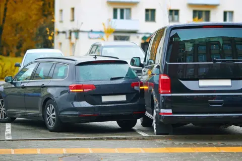 Ciemne kombi i van stoją blisko siebie na parkingu. Widać fragment białego auta. To strefa ruchu, gdzie parkowanie wymaga uwagi.