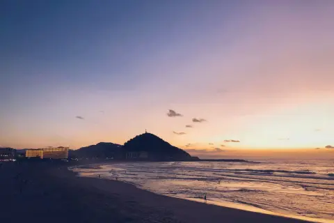 Atardecer en la costa San Sebastián. El cielo se tiñe de tonos rosados y naranjas sobre el mar y la playa, con la silueta del monte Urgull al fondo.