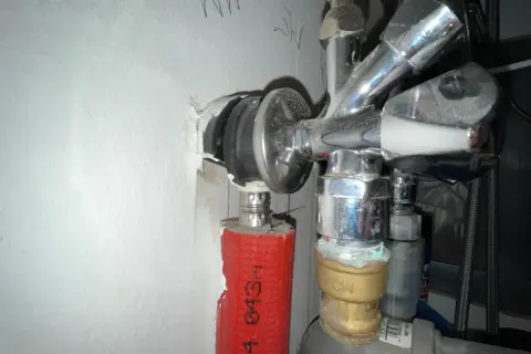 Chromglänzendes Eckventil der Wasserleitung mit rotem Schutzschlauch und sichtbaren Leitungen an einer weißen Wand.