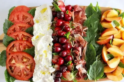 Burrata Antipasti: Tomaten, Burrata, Kirschen, Erdbeeren, Schinken und Pfirsiche auf einem Holzbrett.