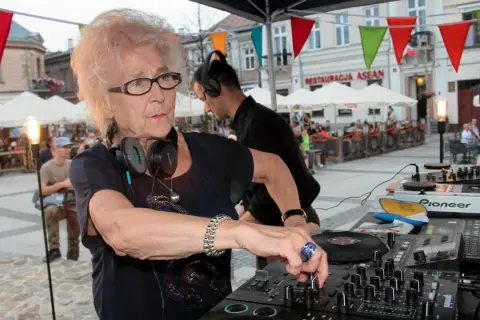 Starsza pani dj z pasją miksuje muzykę na placu miejskim, otoczona ludźmi i kolorowymi flagami.