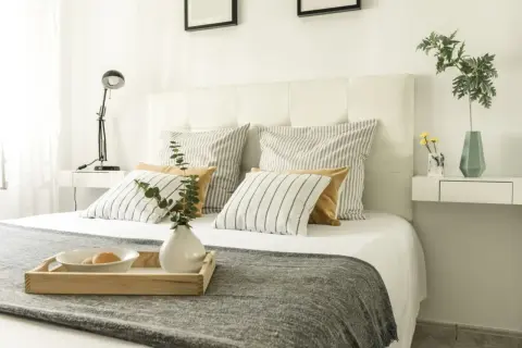 Cama con cabecero blanco, cojines a rayas y mostaza, y bandeja con desayuno. Ideas para como tapizar un cabecero.