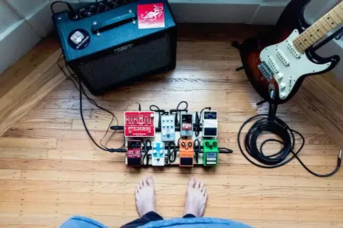 Stojący na drewnianej podłodze pedalboard z efektami gitarowymi, gotowy do gry. W tle wzmacniacz i gitara.