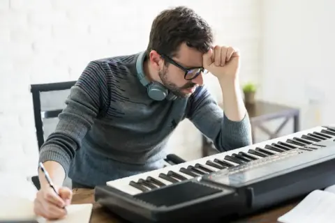 Mężczyzna z okularami i słuchawkami na szyi pochyla się nad klawiaturą pianina, notując coś w zeszycie. Nauka gry na pianinie dla dorosłych bywa wyzwaniem.