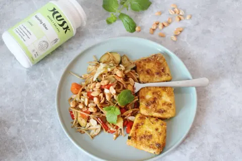 Tallrik med nudlar, grönsaker och friterad tofu. Bredvid en burk HUSK Naturlig fiber, perfekt för mat utan gluten och en balanserad mage.