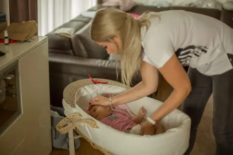 Mama delikatnie kładzie śpiące dziecko do kosza. To pierwszy krok, jak nauczyć dziecko samodzielnego zasypiania.