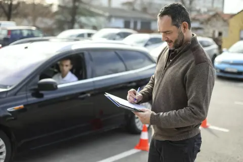 Examinador anotando en una carpeta. Un joven conductor espera en un coche negro. El examen práctico DGT y su precio son importantes para obtener el carnet.