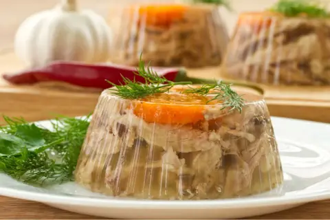Schab po warszawsku w galarecie, ozdobiony marchewką i koperkiem, na talerzu. W tle czosnek i papryczka chili.
