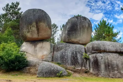 Gigantescas rocas apiladas, como penas de rodas, desafían la gravedad en un paisaje boscoso bajo un cielo azul con nubes.