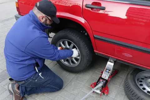 Mężczyzna w niebieskiej kurtce i czapce z daszkiem wykonuje wymianę koła w czerwonym samochodzie, używając podnośnika.