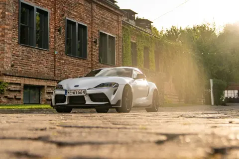 Biała Toyota GR Supra zaparkowana przed ceglanym budynkiem porośniętym bluszczem. Słońce przebija się przez drzewa, tworząc ciepłą, złotą poświatę.