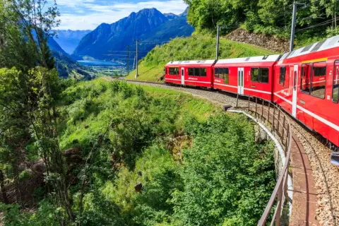 Röda tåget Bernina Express slingrar sig genom ett grönt alplandskap med en sjö och berg i bakgrunden. Undrar du över bernina express pris?